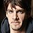 Pixelated static image of a person with light skin tone, dark hair, and neutral expression wearing dark clothing against dark background.