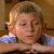 Pixelated static image of a person with light hair resting their head on arms, wearing a blue shirt against a brown background.