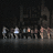 Animated sequence showing a group of stylized figures on a dark stage with reflective floor, transitioning from full lineup to partial view.
