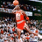A basketball player in a red uniform dribbles while holding an orange ball, with blurred spectators and arena elements visible behind.