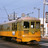 Pixelated image of a yellow and brown tram with dark windows against a light blue sky and urban background.