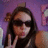 A pixelated image of a person with long dark hair wearing sunglasses and a black top, making a peace sign with their right hand against a purple background with pink and white shapes.