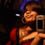 A woman in a red dress and blue headband holds a silver phone, smiling with eyes closed against a dark background.