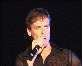 Six distinct performers singing into microphones across various stage settings with different lighting and backgrounds.