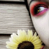 A close-up of a person's eye with red and pink eyeshadow next to a yellow sunflower on a wooden surface.