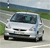 A white sedan with dark-tinted windows is positioned on a road, viewed from a slightly elevated angle, with greenery and a distant structure in the background.
