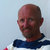 Pixelated portrait of a bald man with light skin wearing a dark blue shirt against a light blue background.