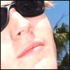Close-up of a person's face wearing dark sunglasses, with light-colored hair visible, against a blurred outdoor background.
