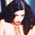 A pixelated close-up of a person with long dark hair, light skin, and red lips, wearing a light-colored top against a blue background.