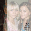 Two people with light-colored hair stand side-by-side against a plain background, one wearing pink and white striped clothing.