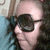A person with dark curly hair and large black sunglasses lies on a blue surface against a patterned background, shown in three