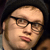 Static pixelated image of a person's face with glasses and dark hair, cropped closely to show upper facial features against a dark background.