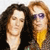 Two people with curly hair stand side-by-side; one wears dark clothing and the other wears a yellow garment with purple sunglasses.