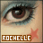 A pixelated close-up of a human eye with brown iris and black eyelashes, below which a brown rectangular banner displays the text "ROCHELLE" in white capital letters.