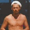 A shirtless man with a white headband holds a water bottle, then pours it into his mouth while standing against a dark background.