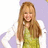 A person with long light brown hair wears a green sleeveless top and white cardigan against a purple background.