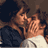 Pixelated static image of two figures in close physical proximity with visible facial features and partial clothing details against a dark background.