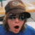 Pixelated static image of a person wearing dark sunglasses and a brown cap, mouth open in an expressive pose against a blurred background.