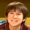 A person with short brown hair and bangs smiles broadly, wearing a maroon shirt against a blurred yellow and brown background.