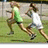 Two figures in athletic wear run on a grassy field with a chain-link fence in the background, accompanied by pink text reading 'nicole' and '#13'.