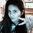 A person with long dark hair and light skin makes a peace sign gesture in front of a blurred background.