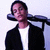 Pixelated static image of a person with dark hair wearing a black jacket over white shirt, arms crossed behind head against light background.