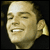 Pixelated static image of a smiling man's face with light skin tone, visible teeth, and partial ear against dark background.