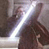 Pixelated static image of a dark figure holding a glowing lightsaber-like object against a muted background with visible dithering and compression artifacts.