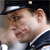 A pixelated close-up of a person wearing a dark cap with a light stripe and a dark jacket with a white collar, looking slightly left.