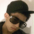A pixelated close-up of a person wearing a black cap and dark sunglasses, with light skin tone visible on the face and ear.