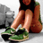 Pixelated image of a person sitting with legs crossed, wearing green and black sneakers, brown shorts, and a dark top against a gray background.