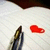 A static image of a pen and a red heart on lined paper with a dark brown background.
