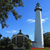 Pixelated static image showing a tall white lighthouse with dark top against blue sky, surrounded by green trees and a smaller dark building.