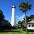 Pixelated static image of a white lighthouse with dark bands near the top, surrounded by green trees and a building under a clear blue sky.