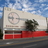 Pixelated static image of a building facade with red accents, a circular emblem, and a tree under a blue sky.