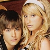 Two people with light brown and blonde hair pose closely together against a dark brown background, one wearing a white headband.