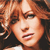Pixelated close-up of a person with light brown curly hair, wearing dark clothing, and a white collar visible at the bottom edge.