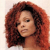 Pixelated close-up of a person with voluminous curly red hair against a light background, showing partial facial features and shoulder.