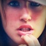 Close-up of a person's face with hand near mouth, visible teeth, and blue eyes in low-resolution static image.