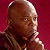 Pixelated close-up of a bald person with brown skin wearing a dark patterned garment against a solid red background, captured in static single-frame format.