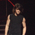 A person with shoulder-length brown hair wears a sleeveless black shirt against a dark background with red lines and horizontal stripes.