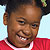 Pixelated close-up of a smiling child with dark skin wearing a red shirt with white trim, dark hair styled in small braids, and a light blue background.