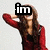 A woman in a red jacket with long brown hair poses with one arm raised behind her head, wearing a black top, against a light gray background.