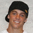 Pixelated close-up of a person with short dark hair wearing a black cap, smiling against a light gray background.