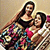 Two figures in a pixelated style, one standing behind the other with arms crossed over chest, both wearing dark tops and colorful patterned lower garments against a light background.