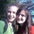 Two people in close proximity, one with light hair and the other with dark hair, against a blurred background with green and blue tones.