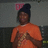 A pixelated figure in an orange shirt with a dark head covering and pink symbol above the head against a gray background.