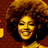 Pixelated static image of a smiling person with voluminous curly hair against a brown background, rendered in warm yellow and orange tones.