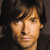 Pixelated close-up of a person with light brown hair and dark eyes, wearing a dark top against a dark background.