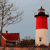 Static pixel art of a red-and-white lighthouse with black trim, a small red-roofed building, and a bare tree against a light blue sky.